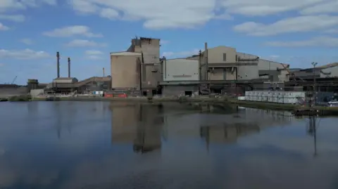 Tata Steel UK An industrial steelmaking site in Port Talbot, South Wales, seen from across a calm body of water. Several large buildings dominate the scene, including tall chimneys and metal-clad structures. Construction materials and equipment are visible near the buildings, suggesting ongoing work. The buildings are reflected clearly in the water, and the sky above is partly cloudy with patches of blue.