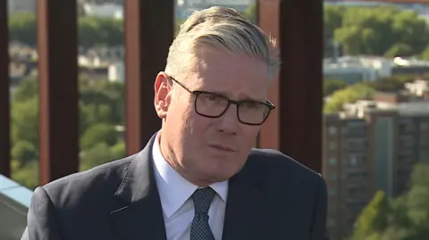 BBC Keir Starmer has short grey hair is smiling in the picture as he wears black square glasses with a black blazer, white shirt and green tie.