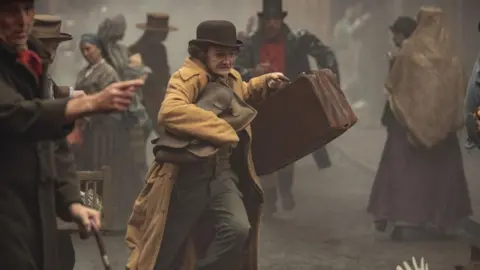 Netflix Jack Gleeson as Byron Hedges wearing a yellow coat and brown cap holding two bags and running in the street