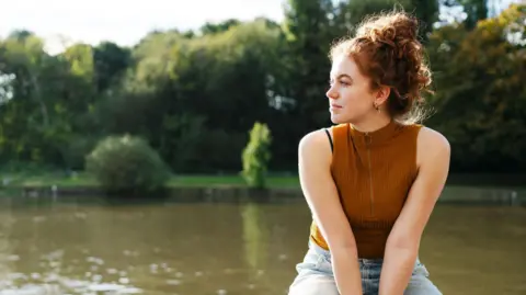Getty Images A woman wearing a brown top and blue jeans with ginger hair tied up, sitting on a ledge with a lake in the background.