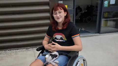 Family handout Wren is smiling into the camera. We see her in her manual wheelchair. She is wearing a blacktop with rainbow edging on the short sleeves and blue denim shorts.  Wren has dark red hair and is wearing glasses.