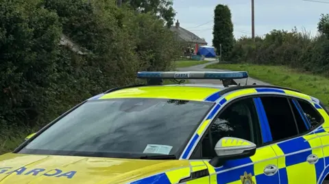 PA Media A blue Garda road closed sign on a country road. Behind it is a police car and cordon. Further back is a building and a blue gazebo outside the building and officers. There are hedges and trees on either side of the road. The sky is cloudy.