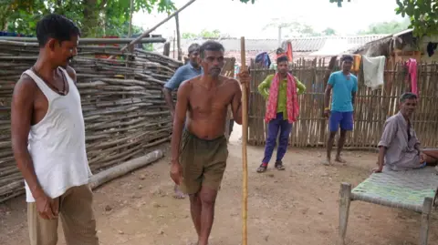 Antariksh Jain Jain/BBC Men standing in a compound in Bastar, Chhattisgarh. The man in the centre is wearing only shorts and is holding a stick while others stand on the sidelines.