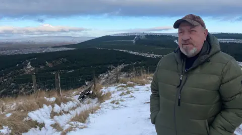 BBC Ian Glenndinning is a man in his 60s wearing a green cap and green coat. He has a closely cropped grey beard and moustache. He's standing on a snowy track with a large expanse of conifer forest behind him under a turquoise cloudy sky