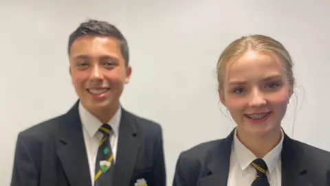 Two pupils in school uniform - which is a black blazer, a black tie with diagonal yellow stripes and a white shirt - standing in front of a plain white wall. The boy on the left, with short dark hair, has a Welsh flag badge on his tie and the girl on the right has blonde hair tied back.