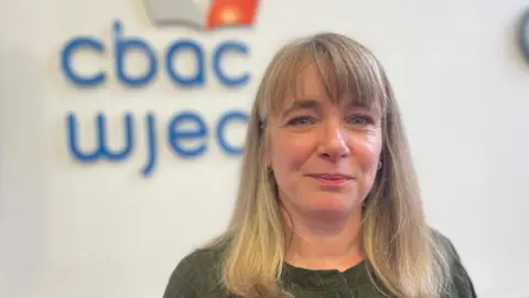 A woman with blonde shoulder length hair with a fringe in front of a wall with a WJEC logo