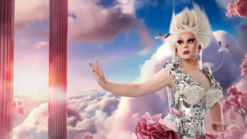Bonnie Ann Clyde. She is wearing a dress with a silver corset and a pink ruffled skirt with a leg slit. She is also wearing a blonde wig which is styled to stick up. She is standing in front of a background of blue skies, green grass and large pink crystals.