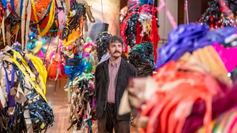 PA Media A man standing in the centre of a large number of suspended colourful sculptures made of various strips of multi-coloured tape and material in Nnena Kalu's installation
