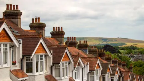 Getty Images A row of rooftops of terraced houses. In the distance there are trees and a hill. The rooftops are red and brown, while the houses are white. The sky is cloudy.