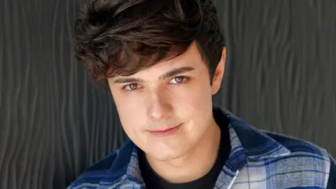 Crunchyroll Zach Aguilar, a youthful-looking man with short black hair, stands in front of a grey background as he smiles into the camera for this studio headshot.