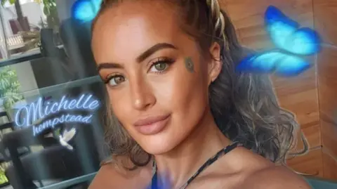 Contributed Michelle Hempstead has dark, curly hair swept back behind her head. This picture is a selfie that has cartoon blue butterflies and her name printed on it. Michelle is smiling with her mouth closed in front of wooden panels.