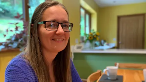 A white woman with brown hair and glasses smiles at the camera wearing a blue cardigan sits at a kitchen table with a cup in the foreground and a vase of sunflowers in the background