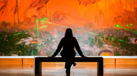 PA Media A silhouetted woman sitting on a gallery bench in front of Mohammed Sami's large painting