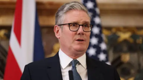 EPA Sir Keir Starmer in a suit and tie standing in front of a blurred Union Jack flag and US flag.