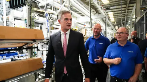PA Media Secretary of State for Business and Trade Peter Kyle MP during a visit to Jaguar Land Rover supplier, Webasto - he is wearing a suit, walking past what appears to be high-tech machinery, with two workers wearing blue branded polo shirts walking next to him