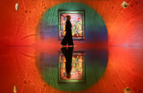 EPA A woman walking past a large colourful painting in a gallery bathed mostly in red light and swirling shapes in Zadie Xa's installation