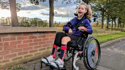 Family handout Wren is looking directly into the camera with a wide smile. She is wearing her school uniform and is sitting in her manual wheelchair.  Wren's hair is tied up in a ponytail and is wearing leg splints and white trainers. The picture has been taken in a wooded area.