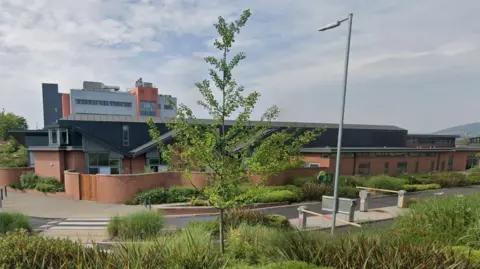 Google A one storey red brick building with a black roof. Belfast City Hospital is visible behind the building. In front of it is bushes and a tree.