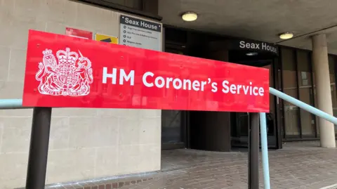 Lewis Adams/BBC A red sign that says HM Coroner's Service. It is at the entrance to Essex Coroner's Court, which is a cream building that has a black revolving door and brick paving outside.