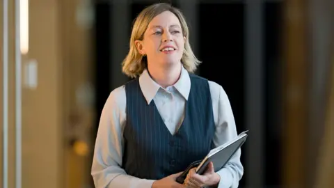 PA Media Jenny Gilruth walking inside the Scottish Parliament, carrying a black pad. She has shoulder-length blone hair and is wearing a pinstriped waistcoat and a light blue shirt.